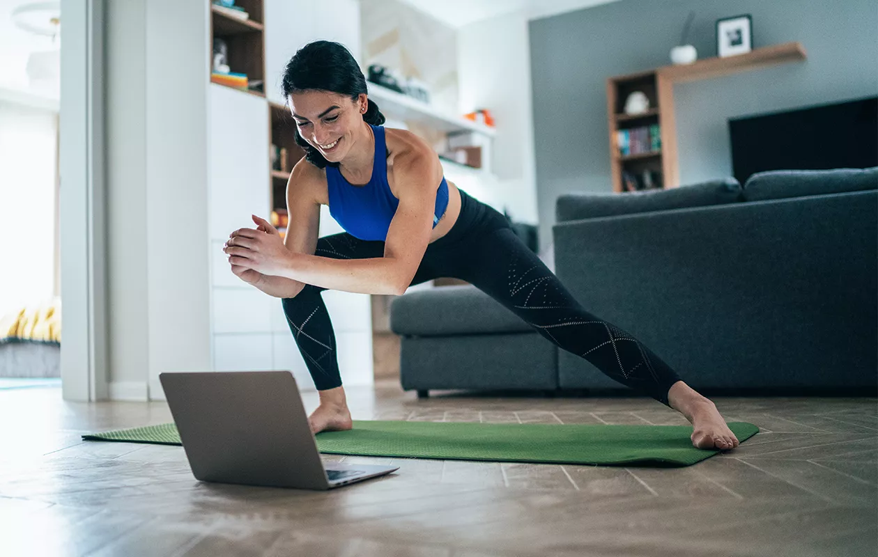Nainen venyttelee joogamatolla kannettavan tietokoneen edessä. Tämä pakaratreeni kotona saa taatusti hien pintaan.