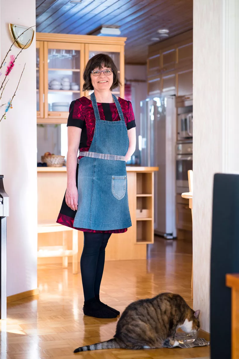 Kirsi Peltola seisoo keittiössään esiliina yllään. Hänen jaloissaan kissa juo vettä kupista. 