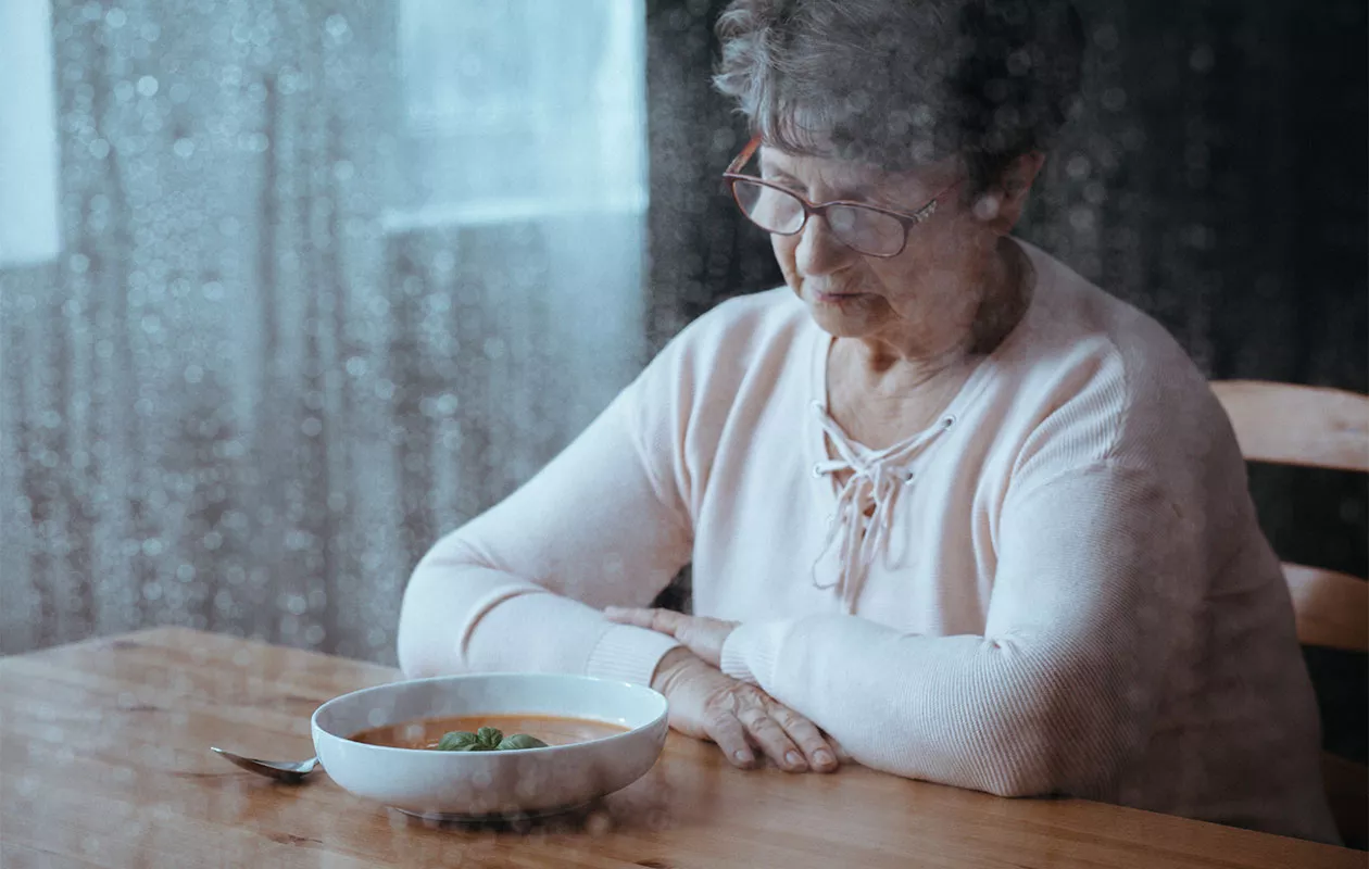 Sateisen ikkunan takaa otettu kuva vanhemmasta naisesta, joka katsoo keittolautasta. Aikuisten syömishäiriöt voivat koskettaa minkä ikäisiä ihmisiä tahansa.