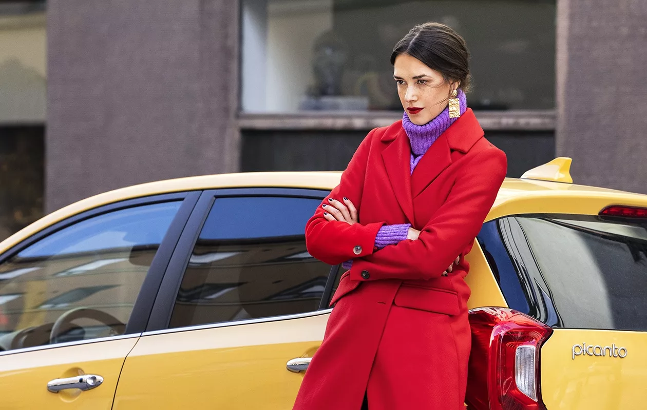 Nainen nojaa taksiin punainen villakangastakki yllään.